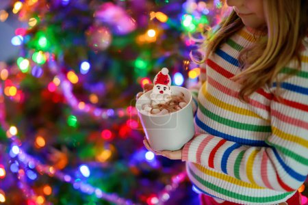 Foto de Niña sosteniendo taza con chocolate caliente con malvaviscos como Santa Claus. Niño cerca del árbol de Navidad decorado con luces - Imagen libre de derechos
