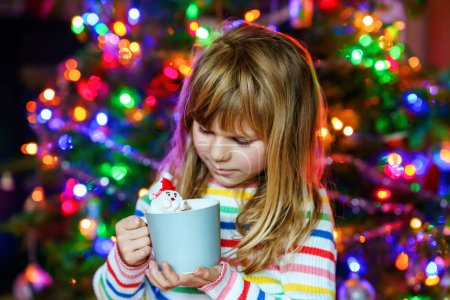 Téléchargez les photos : Petite fille tenant une tasse avec du chocolat chaud avec des guimauves comme le Père Noël. Enfant près de l'arbre de Noël décoré de lumières - en image libre de droit