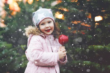 Téléchargez les photos : Jolie petite fille maternelle souriante sur le marché de Noël allemand. Enfant heureux en vêtements d'hiver mangeant doux sucré pomme de Noël glacée avec des lumières sur le fond. Famille, tradition, concept de célébration - en image libre de droit