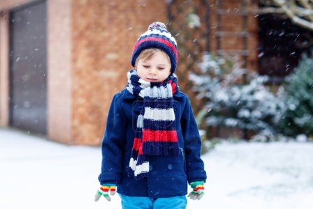 Téléchargez les photos : Mignon petit enfant drôle dans des vêtements colorés de mode d'hiver s'amuser et jouer avec la neige, à l'extérieur pendant les chutes de neige. Loisirs en plein air actifs avec enfants. Enfant garçon et tout-petit attrapant flocons de neige - en image libre de droit