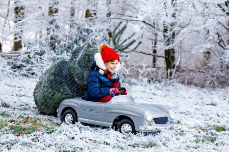 Téléchargez les photos : Heureuse petite fille souriante conduisant une voiture jouet avec arbre de Noël. Drôle d'enfant d'âge préscolaire en vêtements d'hiver apportant arbre de Noël taillé de forêt enneigée. Famille, tradition, vacances - en image libre de droit