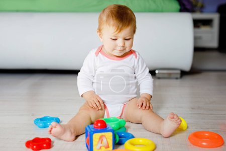 Photo for Adorable baby girl playing with educational toys in nursery. Happy healthy child having fun with colorful different toys at home. Kid trying to build plastic pyramid and using blocks with letters. - Royalty Free Image