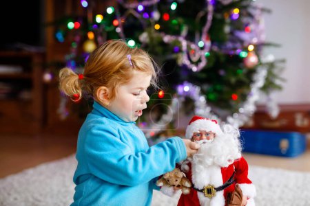 Photo for Adorable toddler girl playing with gifts and Christmas Santa Claus toys. Little child having fun with decorated and illuminated Xmas tree with lights on background. Happy healthy funny baby girl - Royalty Free Image
