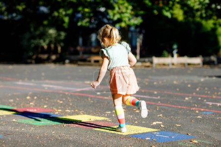 Téléchargez les photos : Mignon petit tout-petit fille jouer jeu de marelle dessinée avec des craies colorées sur l'asphalte. Petit enfant actif sautant sur l'aire de jeux à l'extérieur par une journée ensoleillée. Activités estivales pour les enfants - en image libre de droit
