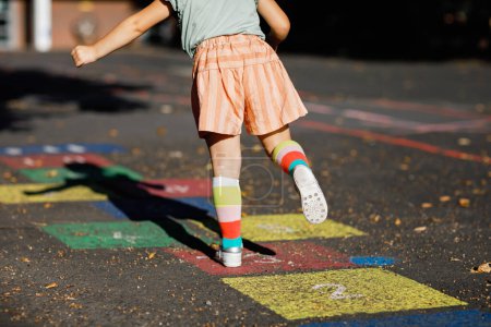 Téléchargez les photos : Gros plan de jambes de petite fille tout-petit jouant jeu de marelle dessinée avec des craies colorées sur l'asphalte. Petit enfant actif sautant sur l'aire de jeux à l'extérieur par une journée ensoleillée. Activités estivales pour les enfants - en image libre de droit