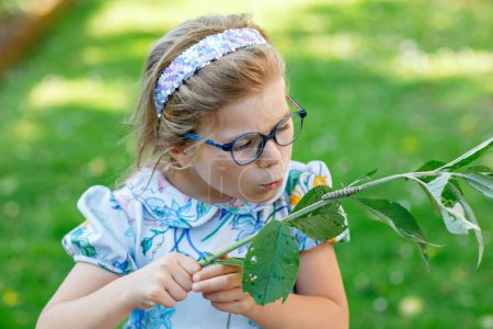 Téléchargez les photos : Petite fille préscolaire regarde chenille grimper sur la plante. Heureux enfant excité regarder et apprendre les insectes dans les jardins domestiques - en image libre de droit