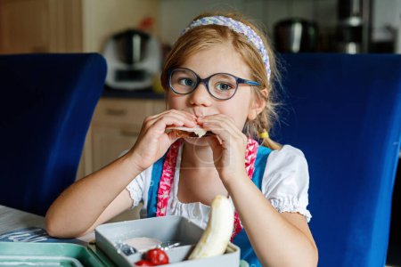 Foto de Linda niña con anteojos comiendo sándwich y frutas durante el descanso entre clases. Alimento saludable para niños. Desayuno lonchera para niños en la escuela - Imagen libre de derechos
