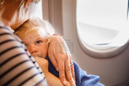 Foto de Mujer viajando con un niño pequeño en avión. Triste niña cansada sentada con mamá por la ventana del avión. Concepto de maternidad. Bebé llorón - Imagen libre de derechos