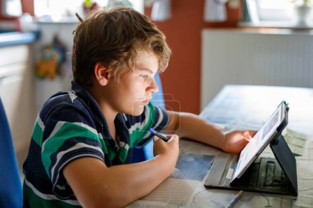Téléchargez les photos : Préado garçon apprenant à la maison sur ordinateur portable pour l'école. Un écolier faisant ses devoirs et utilisant un cahier et des gadgets modernes. Concept d'enseignement à domicile - en image libre de droit