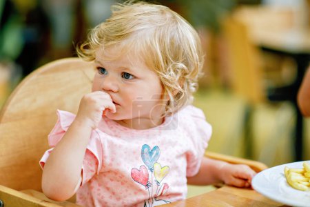 Photo for Adorable toddler girl eating healthy vegetables and unhealthy french fries potatoes. Cute happy baby child taking food from dish at daycare or nursery canteen - Royalty Free Image