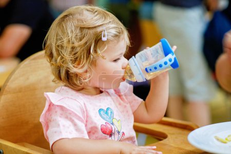 Photo for Adorable toddler girl drinking formula milk or water from bottle. Cute happy baby child taking food from dish at daycare or nursery canteen - Royalty Free Image