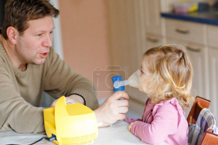 Photo for Little toddler girl making inhalation with nebulizer at home. Father helping and holding the device. Child having flu, cough and bronchitis. asthma inhaler inhalation steam sick concept. - Royalty Free Image