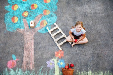 Téléchargez les photos : Jolie peinture de petite fille avec des craies colorées récolte de pommes de pommier sur asphalte. Mignon enfant d'âge préscolaire avec s'amuser avec une image à la craie. Loisirs créatifs pour les enfants, dessin et peinture - en image libre de droit