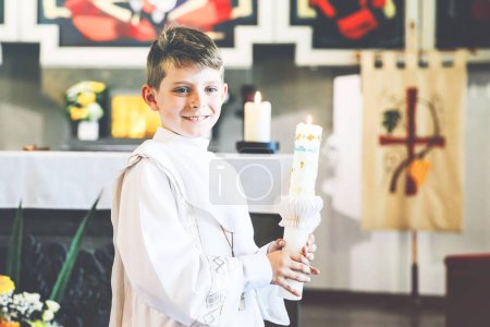 Téléchargez les photos : Petit garçon recevant sa première sainte communion. Joyeux enfant tenant une bougie du baptême. Tradition en curch catholique. Enfant en robe traditionnelle blanche dans une église près de l'autel - en image libre de droit