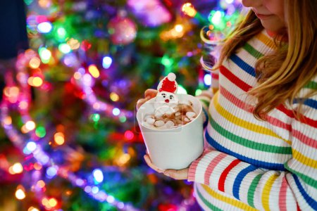 Téléchargez les photos : Petite fille tenant une tasse avec du chocolat chaud avec des guimauves comme le Père Noël. Enfant près de l'arbre de Noël décoré de lumières - en image libre de droit