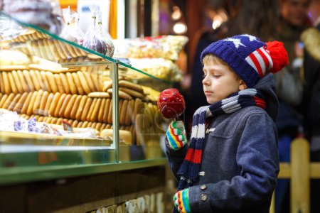 Téléchargez les photos : Petit garçon mignon près de stand doux avec du pain d'épice et des noix. Joyeux enfant mangeant sur une pomme couverte de sucre rouge. Doux traditionnel sur le marché de Noël allemand. - en image libre de droit