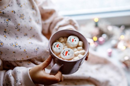 Foto de Niña irreconocible sosteniendo taza con chocolate caliente con malvaviscos como muñeco de nieve. Niño sentado cerca de la ventana decorada de Navidad con luces - Imagen libre de derechos