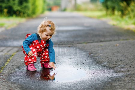 Photo for Little toddler girl wearing rain boots and trousers and walking during sleet, rain on cold day. Baby child having fun with jumping in a puddle. Active outdoors leisure and activity with little kids - Royalty Free Image