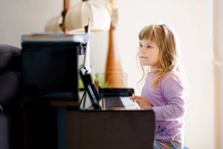 Photo for Beautiful little toddler girl playing piano in living room. Cute preschool child having fun with learning to play music instrument. Early musical education for children - Royalty Free Image