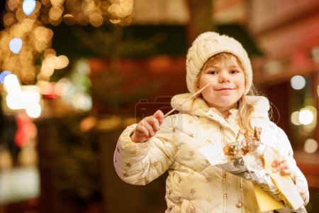 Foto de Niña preescolar, linda niña comiendo churros dulces cubiertos de chocolate con decoración y luces en el fondo. Niño feliz en el mercado de Navidad en Alemania - Imagen libre de derechos