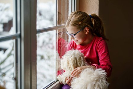 Foto de Niña sentada junto a la ventana con su perro mascota maltés en casa. Niño feliz y lindo cachorro mirando hacia fuera en el paisaje de nieve de invierno. Amor, amistad, familia animal - Imagen libre de derechos