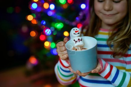Photo for Little child girl holding cup with hot chocolate with marshmallows as snowman. Kid sitting near Christmas tree decorated with lights - Royalty Free Image