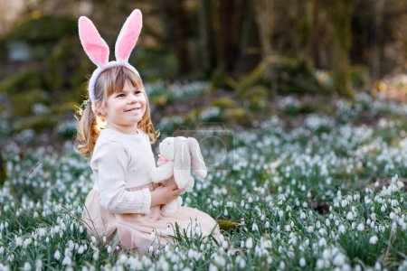 Foto de Adorable niña con orejas de conejo de Pascua sosteniendo un juguete de felpa suave en el bosque de primavera en un día soleado, al aire libre. Lindo niño feliz con un montón de flores de nieve. Primavera, concepto de vacaciones cristianas - Imagen libre de derechos