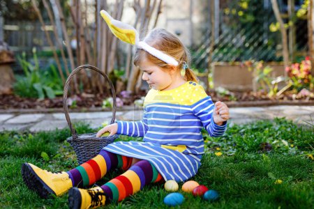 Photo for Beautiful funny little toddler girl with bunny ears having fun with traditional Easter eggs hunt on sunny day, outdoors. Happy child celebrating holiday. Baby find and collect colored eggs in garden. - Royalty Free Image