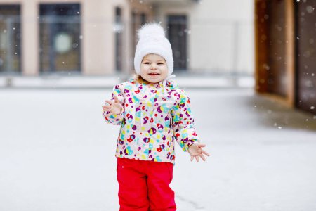Téléchargez les photos : Adorable petite fille en bas âge marchant dehors en hiver. Bébé mignon mangeant des bonbons sucrés sucrés. Enfant qui s'amuse par temps froid. Porter des vêtements chauds et colorés bébé et chapeau avec des bobbles - en image libre de droit