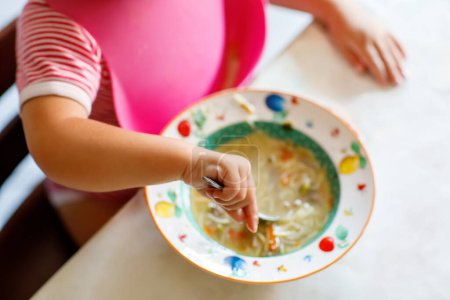 Photo for Adorable baby girl eating from spoon vegetable noodle soup. Healthy food, child, feeding and development concept. Cute toddler child with spoon sitting in highchair and learning to eat by itself. - Royalty Free Image