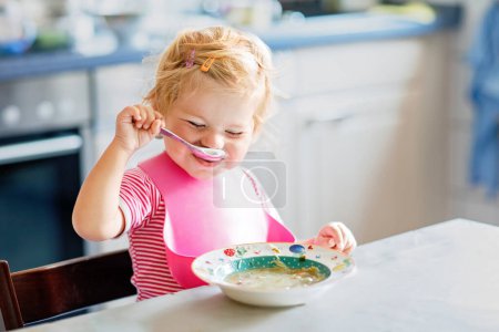Photo for Adorable baby girl eating from spoon vegetable noodle soup. Healthy food, child, feeding and development concept. Cute toddler child with spoon sitting in highchair and learning to eat by itself. - Royalty Free Image
