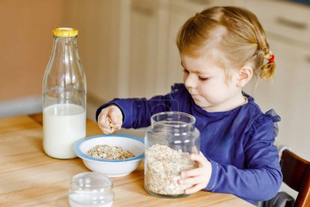 Photo for Adorable toddler girl eating healthy oatmeals with milk for breakfast. Cute happy baby child in colorful clothes sitting in kitchen and having fun with preparing oats, cereals. Indoors at home. - Royalty Free Image