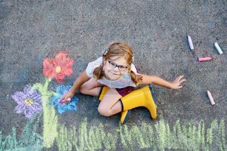 Foto de Linda niña y flores pintadas con tiza de colores sobre asfalto. Feliz niño preescolar divirtiéndose con la pintura de tiza. Ocio creativo para niños, dibujo y pintura - Imagen libre de derechos