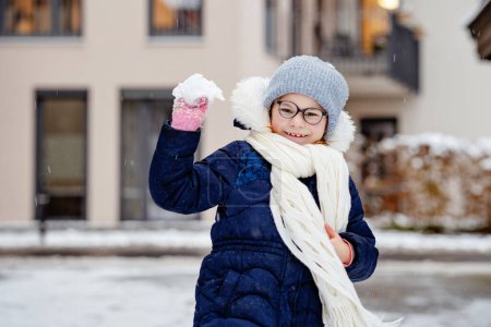 Foto de Retrato de adorable niña al aire libre en el frío día de invierno. Lindo niño preescolar en ropa de abrigo, con sombrero de punto y bufanda. Chica feliz con gafas que se divierten con la nieve - Imagen libre de derechos