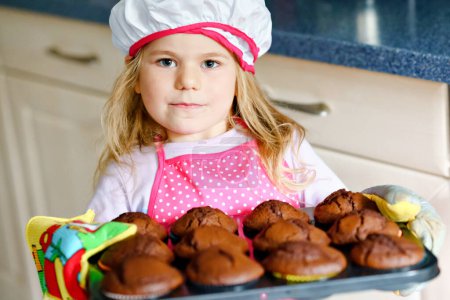 Téléchargez les photos : Petite fille mignonne tout-petit tenant des muffins au chocolat frais cuits à la maison cupcakes à la maison à l'intérieur. Adorable enfant blond préscolaire avec tablier avec lapin et biscuit à la carotte dans la cuisine domestique - en image libre de droit