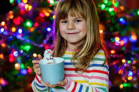 Téléchargez les photos : Petite enfant fille tenant tasse avec chocolat chaud avec guimauves comme bonhomme de neige. Enfant assis près de l'arbre de Noël décoré de lumières - en image libre de droit