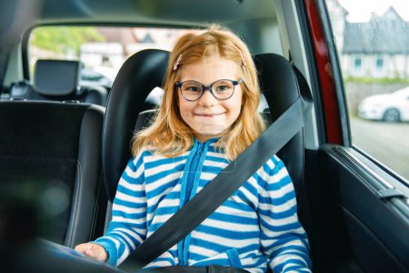 Téléchargez les photos : Petite fille d'âge préscolaire assise dans son siège auto. Happy Child avec des lunettes de vue Lire un livre, sourire sur le chemin des vacances en famille pendant les embouteillages - en image libre de droit