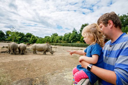 Téléchargez les photos : Mignon adorable tout-petit fille et père regardant rhinocéros sauvages dans le zoo. Bébé enfant heureux, fille et papa, famille s'amuser avec les animaux safari parc sur chaude journée d'été. Irlande. - en image libre de droit