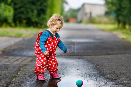 Photo for Little toddler girl wearing rain boots and trousers and walking during sleet, rain on cold day. Baby child having fun with jumping in a puddle. Active outdoors leisure and activity with little kids - Royalty Free Image