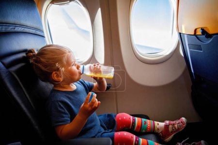 Foto de Adorable niñita viajando en un avión. Niño pequeño bebiendo jugo de naranja sentado cerca de la ventana del avión. Viajar al extranjero con niños. Familia en vacaciones de verano - Imagen libre de derechos