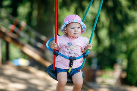 Photo for Cute adorable toddler girl swinging on outdoor playground. Happy smiling baby child sitting in chain swing. Active baby on sunny summer day outside. Family leisure and activity outdoors - Royalty Free Image