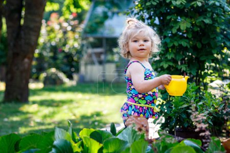 Foto de Linda niña en traje de baño colorido regando plantas y flores en flor en el jardín doméstico en el día caluroso de verano. Adorable niño pequeño que se divierte jugando con agua y puede
. - Imagen libre de derechos