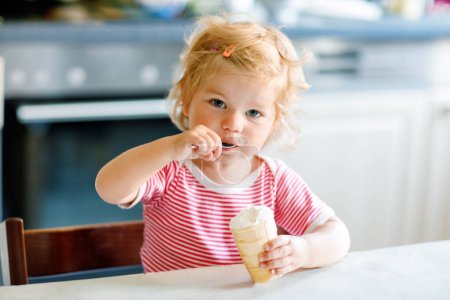 Photo for Adorable baby girl eating from spoon sweet ice cream in waffle cone. food, child, feeding and development concept. Cute toddler, daughter with spoon sitting in highchair and learning eat by itself. - Royalty Free Image