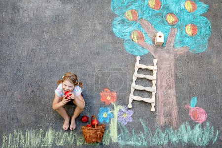 Téléchargez les photos : Jolie peinture de petite fille avec des craies colorées récolte de pommes de pommier sur asphalte. Mignon enfant d'âge préscolaire avec s'amuser avec une image à la craie. Loisirs créatifs pour les enfants, dessin et peinture - en image libre de droit