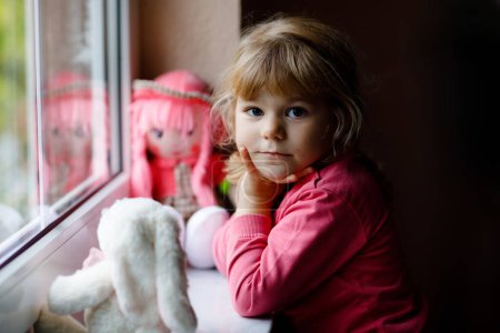 Photo for Cute toddler girl sitting by window and looking out on rainy day. Dreaming child with doll and soft toy feeling happy. Self isolation concept during corona virus pandemic time. Lonely kid - Royalty Free Image