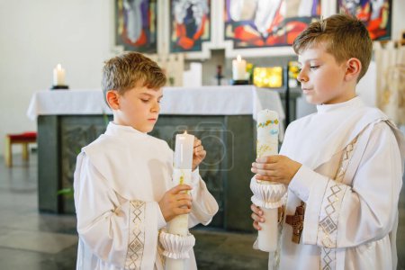 Téléchargez les photos : Deux petits garçons recevant sa première sainte communion. Des enfants heureux tenant une bougie du baptême. Tradition en curch catholique. Des enfants dans une église près de l'autel. Frères et sœurs, frères et sœurs en robes blanches - en image libre de droit