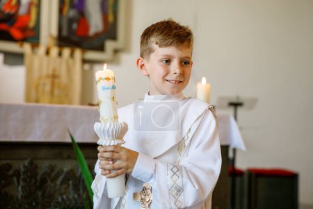 Téléchargez les photos : Petit garçon recevant sa première sainte communion. Joyeux enfant tenant une bougie du baptême. Tradition en curch catholique. Enfant en robe traditionnelle blanche dans une église près de l'autel - en image libre de droit
