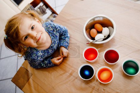 Foto de Emocionada niña para colorear huevos para Pascua. Lindo niño feliz buscando sorprendido en coloridos huevos de colores, celebrando la fiesta con la familia. Desde arriba, cara no reconocida - Imagen libre de derechos