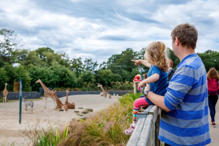 Téléchargez les photos : Mignon adorable tout-petit fille et père regarder et nourrir girafe dans zoo. Bébé enfant heureux, fille et papa, famille s'amuser avec les animaux safari parc sur chaude journée d'été. Irlande. - en image libre de droit