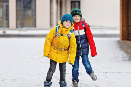 Téléchargez les photos : Deux petits garçons de classe primaire marchant jusqu'à l'école pendant les chutes de neige. Enfants heureux s'amuser et jouer avec la première neige. Frères et sœurs et meilleurs amis avec sac à dos en vêtements d'hiver colorés - en image libre de droit